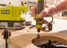 Hand operating a rotary tool to file cut-out in a piece of wood that is clamped to a work bench.