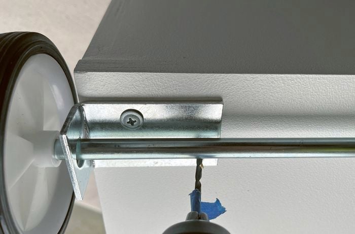 A white wheel and a metal axle attached to a white piece of wood with a metal bracket.