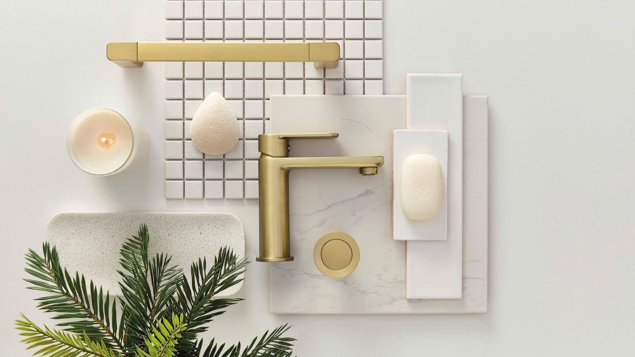 Flatlay image of bathroom fixtures including a gold basin mixer tap, gold hand towel rail and soap dish next to a candle and a white marble tile