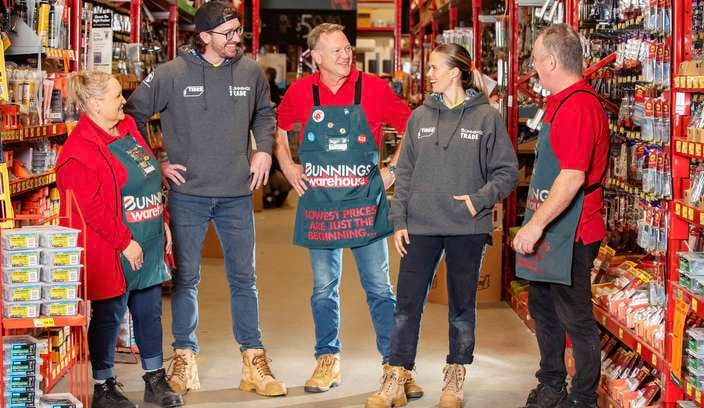 Five Bunnings team members standing together in a store aisle, talking amongst themselves stocked with hardware products.