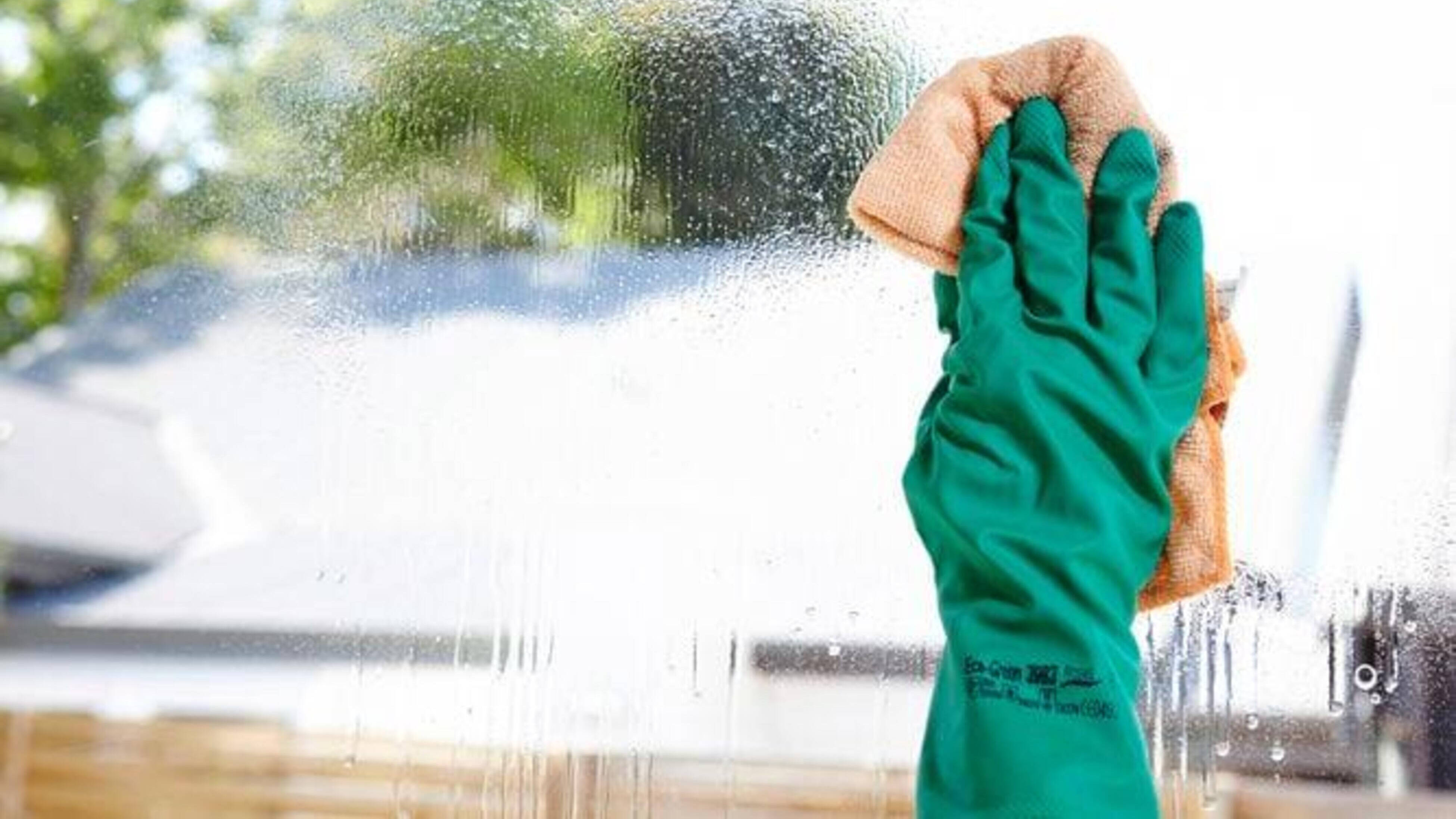 A green gloved hand holding a microfibre cloth wipes a freshly sprayed window.