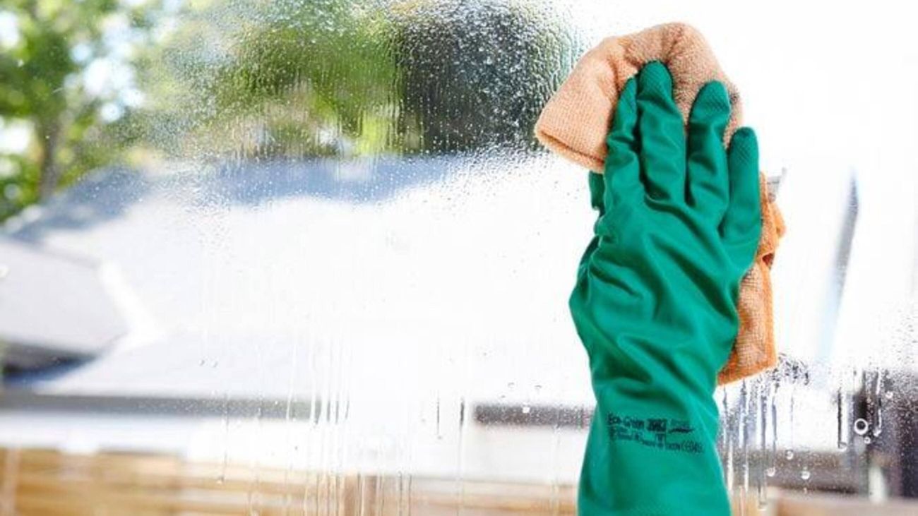 A green gloved hand holding a microfibre cloth wipes a freshly sprayed window.