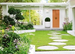 A well-manicured garden with green grass, white stepping stones, white edging, green trees and blooming flowers