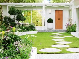 A well-manicured garden with green grass, white stepping stones, white edging, green trees and blooming flowers