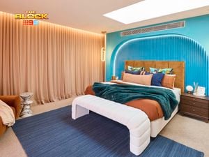 A modern bedroom featuring a large bed with white and brown bedding, colorful pillows, and a green throw blanket. The blue headboard wall has an arched design and pendant lights on either side. A wooden nightstand with decorative items sits next to the bed. The room includes a blue rug, beige curtains, and a rectangular skylight in the ceiling. 