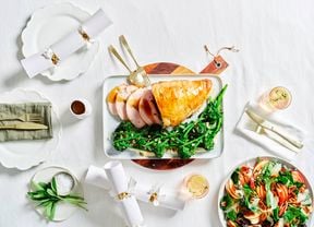 Image of a main dish of Turkey Roulade with broccolini and beans with a zesty dressing, a side dish of fresh salad on a white linen table setting with cutlery and drinks.