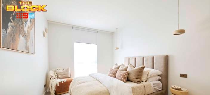 A furnished bedroom with a bed, carpet, bedside table, and chair included in it and The Block logo included on the image.