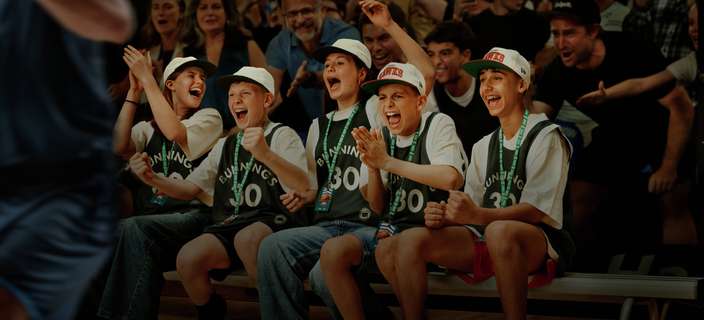 Fans wearing matching jerseys and hats sit on a bench in a crowded basketball arena, clapping and cheering as a player runs past in the foreground.