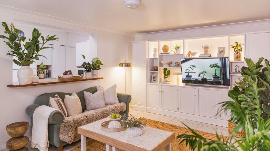 Wide view of a styled living room featuring a green sofa, coffee table, indoor plants, and white built‑in cabinetry with a television.