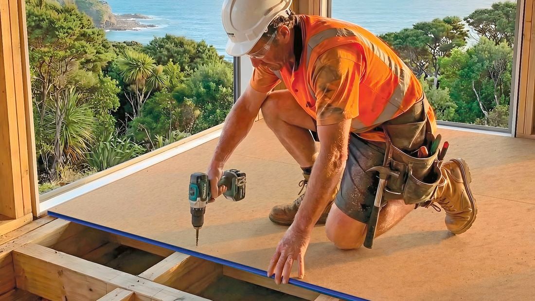 A construction worker in a hard hat and high-visibility vest kneels on timber sub flooring, using a power drill to install a floor panel inside a house overlooking the coast.