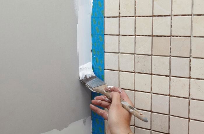 A person painting down the edge of a plasterboard wall next to a tiled wall with masking tape
