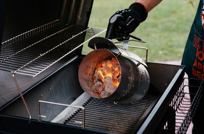 Person pouring hot charcoal briquettes into a smoker 