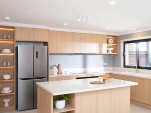 A bright, modern kitchen with natural sunlight features light wood cabinetry, a large island with white countertops, and a window