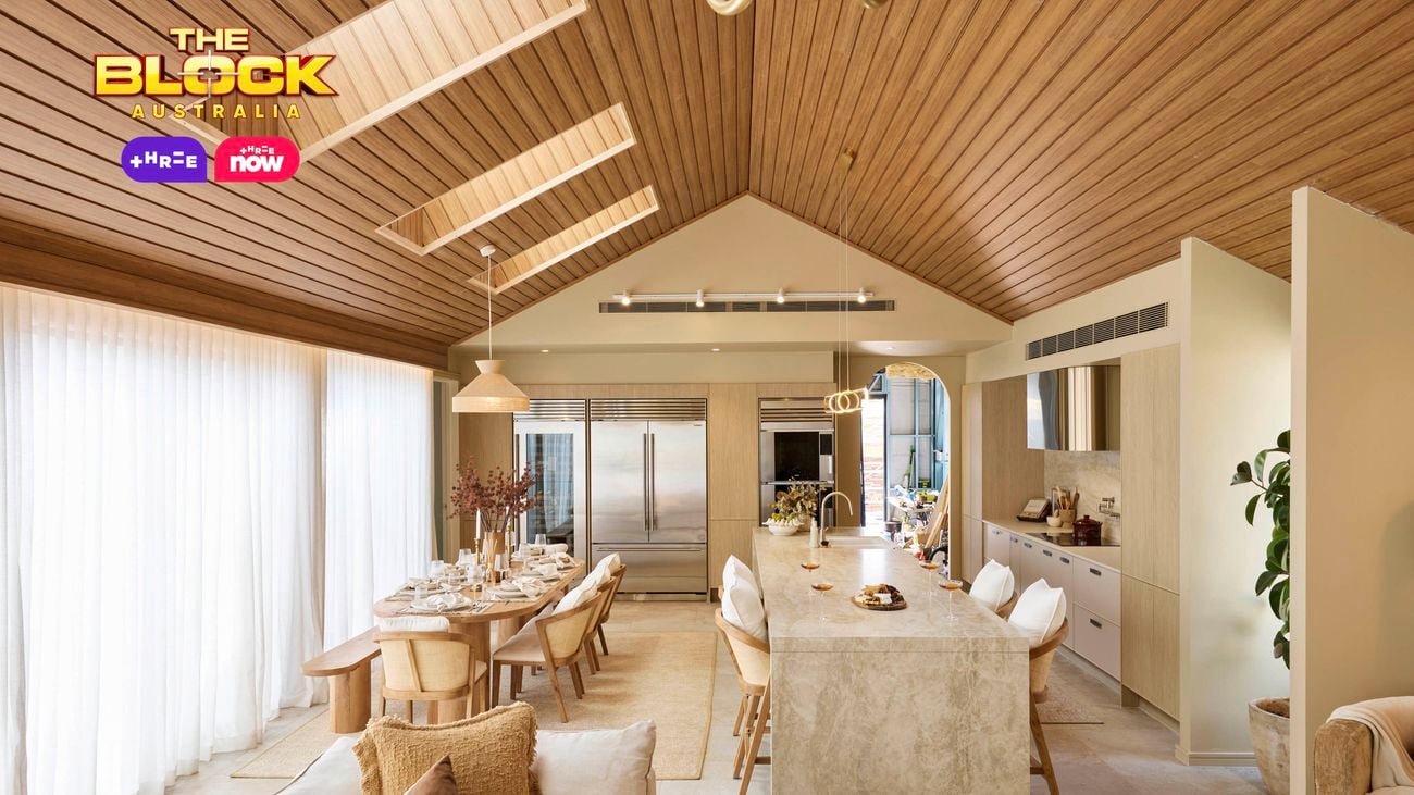 Open-plan kitchen and dining space from The Block, featuring a vaulted timber ceiling, marble island with bar stools, large fridge, neutral-toned dining area, and floor-to-ceiling curtains.