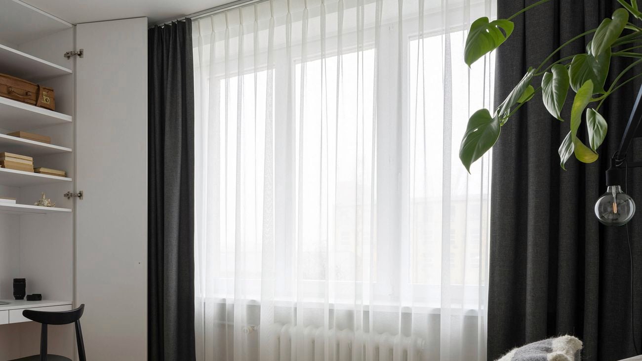 A close-up of light-coloured curtains in a bedroom with a second set of energy saving blockout curtains, a leafy green plant, and an industrial-style lamp and single lightbulb, an open cupboard with shelving containing books, a light brown suitcase and knick knacks, a built-in desk, and a chair