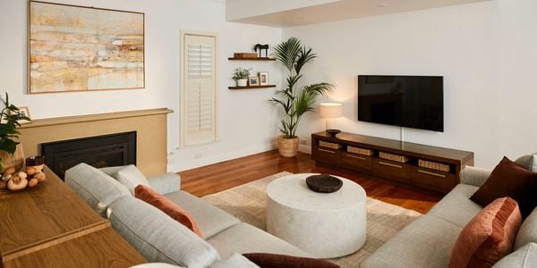 Light-filled living room with wooden floors, a wall-mounted TV above a low timber cabinet with baskets, a round coffee table on a rug, neutral sofas, indoor plants, and minimalist wall shelves.