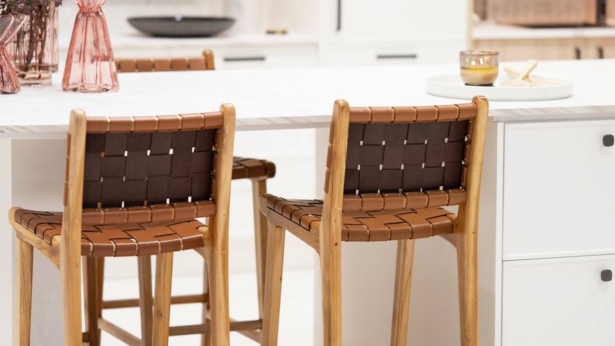Contemporary kitchen display featuring a white island bench, woven bar stools, integrated shelving and warm ambient lighting.