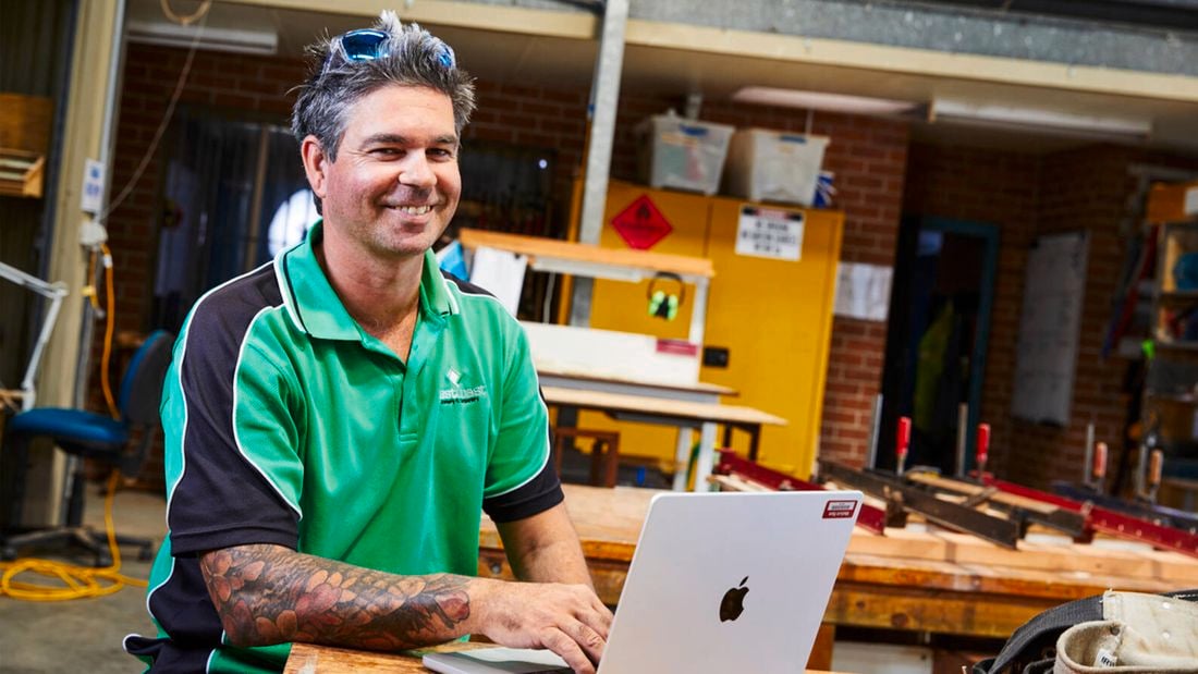 A smiling man in a green and black polo shirt with tattoos is working on a laptop in a workshop setting.]