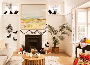 A living room decorated for Halloween with black bat cut-outs on the walls, pumpkins, candles and a table with orange jack-o'-lantern buckets. A sofa, fireplace and a large painting are also visible