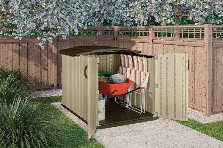 Corner of a fenced garden with small storage cabinet containing a wheelbarrow and folding chairs
