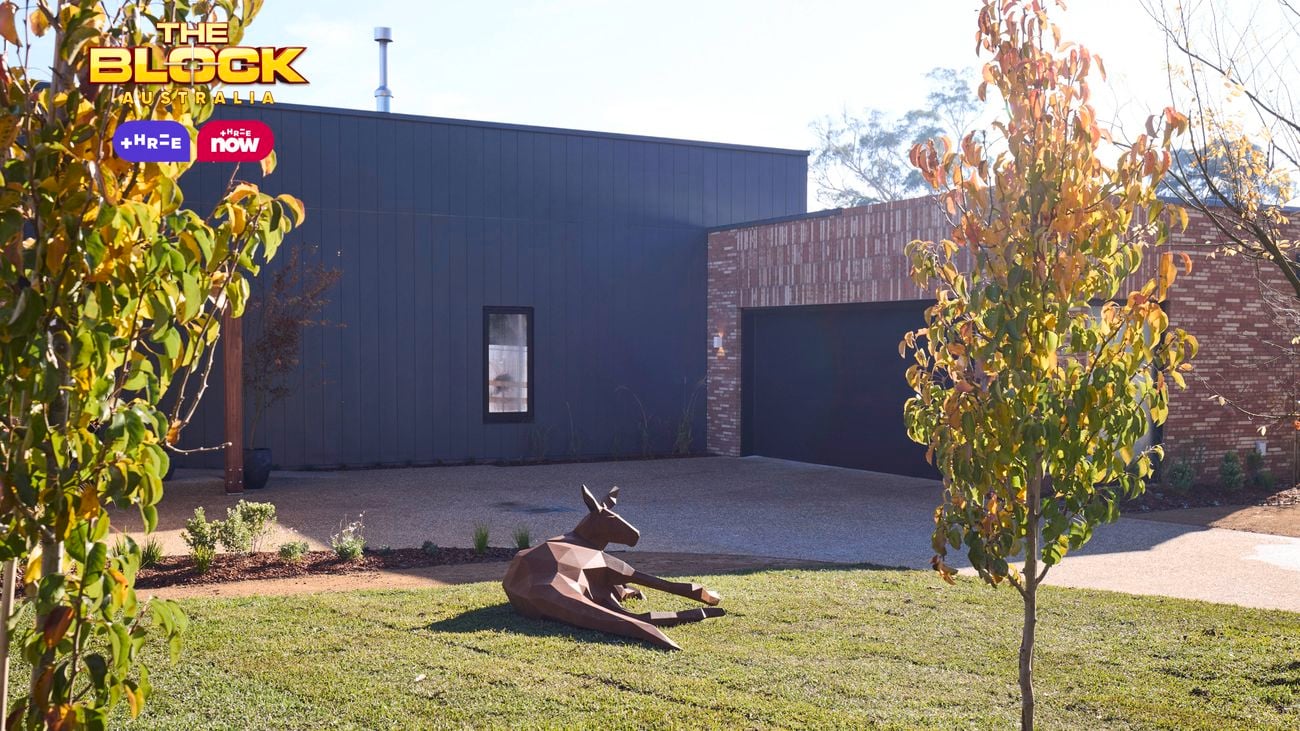 A kangaroo made out of steel laying down on a piece of grass with trees and a driveway at the front of a house.
