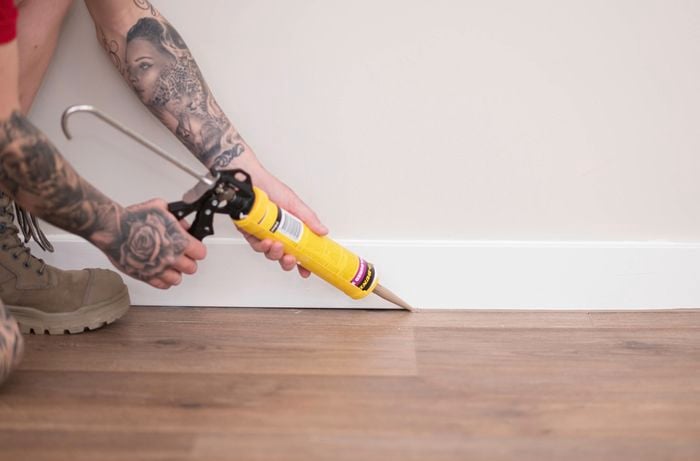 A person is applying a bead of silicone along the bottom edge of the skirting board using a caulking gun. 