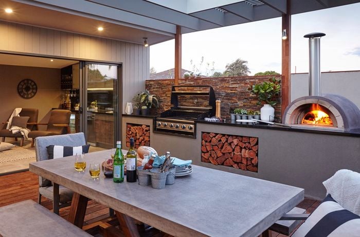 An outdoor room with wood-fired pizza oven, BBQ and furniture looking into a living room