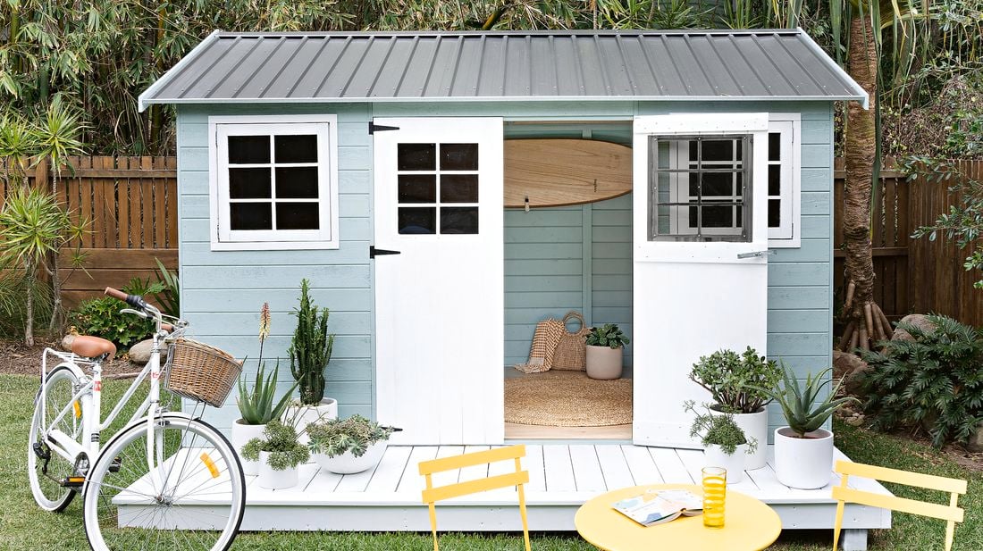 A painted timber garden shed with a bike and garden furniture in a backyard