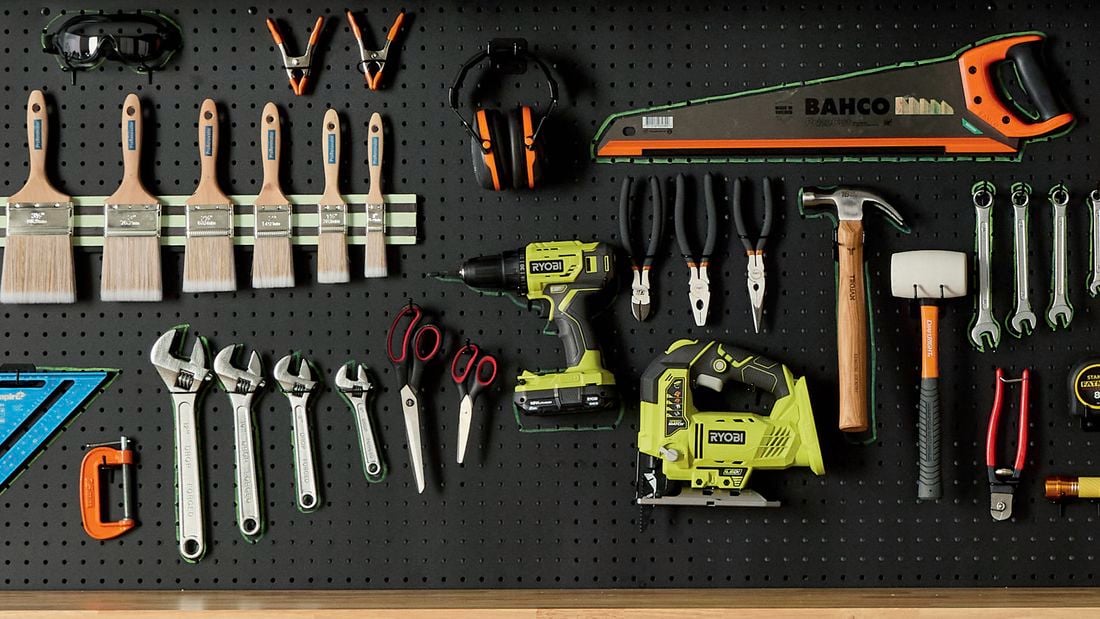 A pegboard displaying a variety of hand and power tools, including paint brushes, wrenches, pliers, a saw, a drill, and a jigsaw.