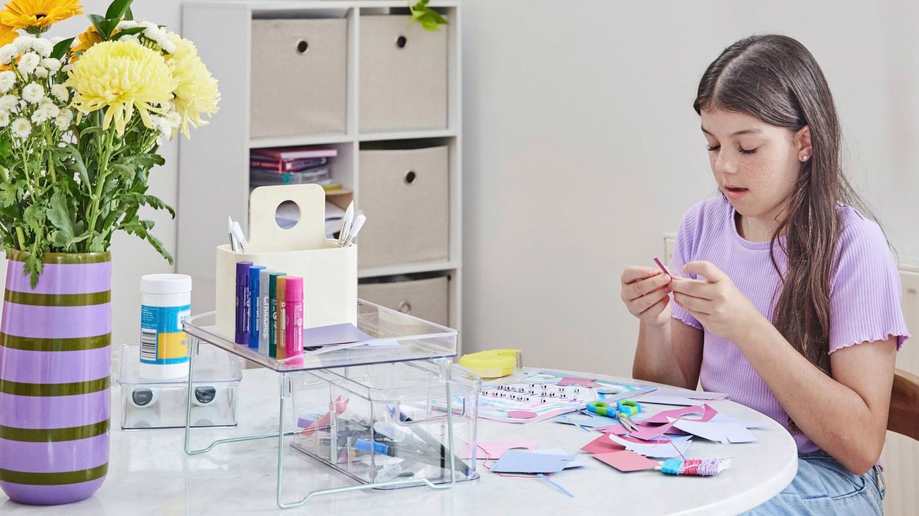 Young girl focused on a craft project at a round white desk. A tall purple vase with black stripes and colourful flowers, storage organiser with craft materials and cube storage against one wall.