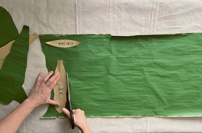 Hands cutting around a leaf template on green-painted kraft paper using scissors with a Sharpie nearby.