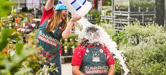 Two Bunnings Warehouse staff members in store uniforms conduct an ice bucket challenge inside a garden centre, with one person seated on a chair while another pours a bucket of water over them, surrounded by potted plants.