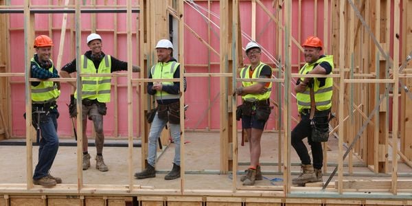 An image of five tradies wearing yellow hi-vis - two in orange hard hats and three in white hard hats - looking to camera through timber frames on a construction site