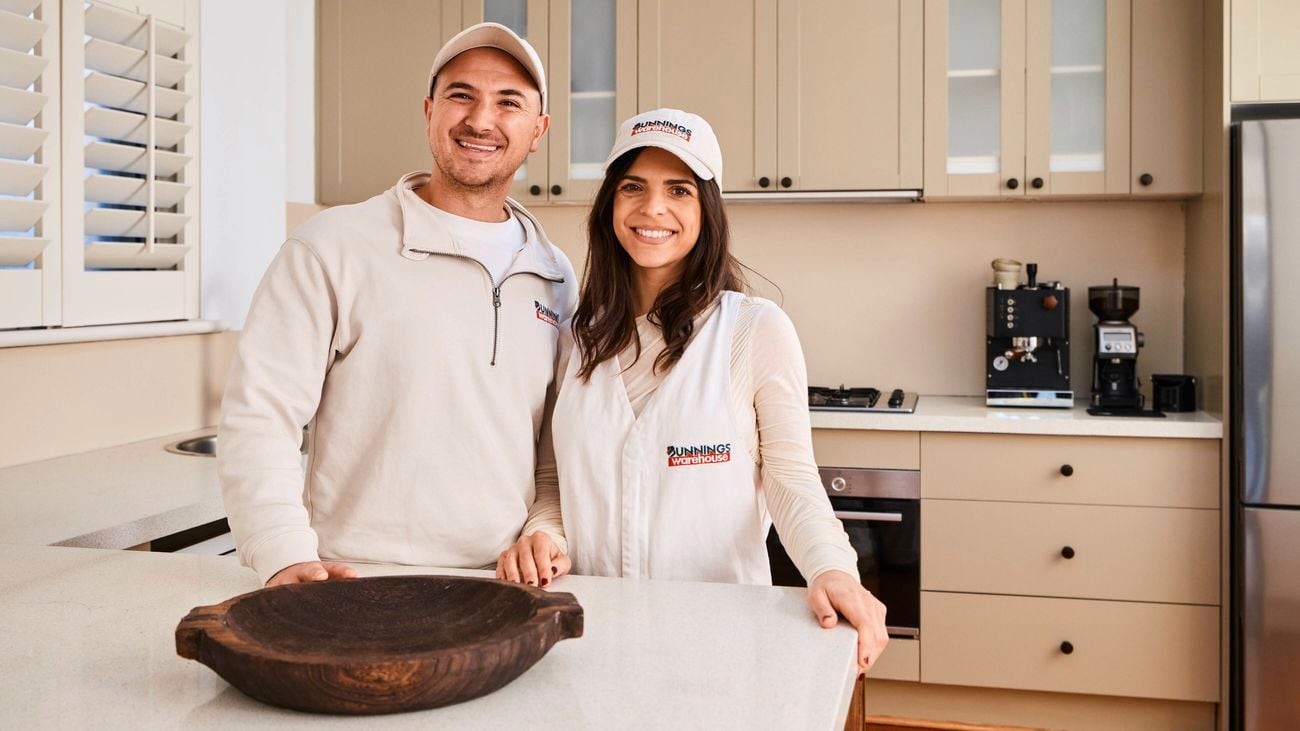   A couple consists of a man and a woman standing side by side behind a kitchen island in a modern beige kitchen, both wearing branded caps and casual tops, with cabinets, a cooktop, coffee machine, and stainless steel fridge visible in the background.