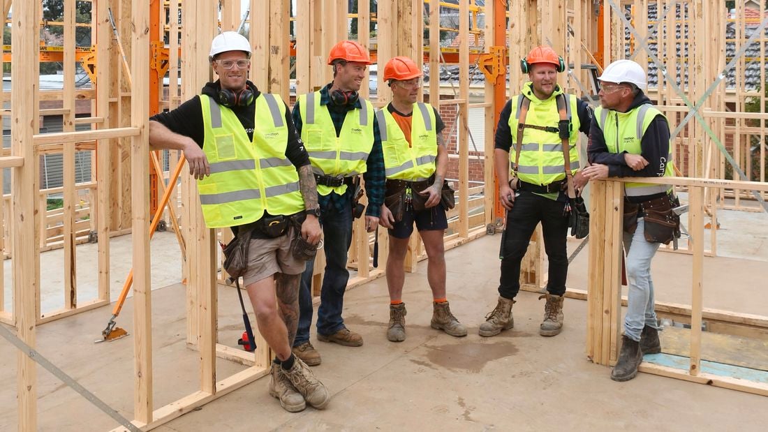 Five carpenters, two in orange hard hats and three in white hard hats, stand amongst frame and truss work onsite
