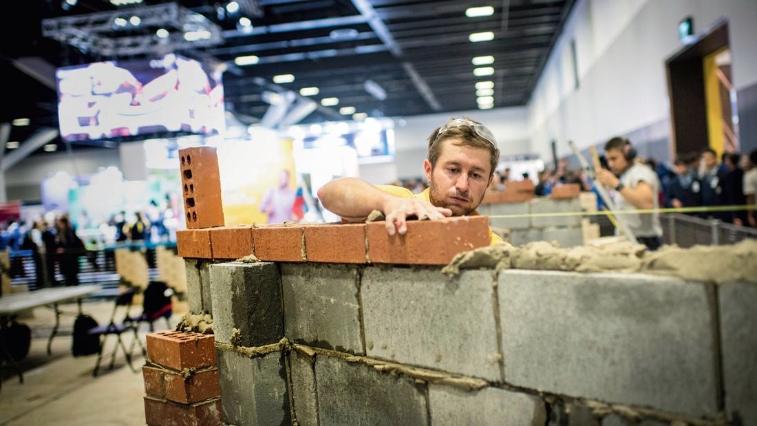 A person places a red brick onto a partially built brick and concrete block wall inside a large indoor exhibition hall. The wall is mid-construction with visible mortar, and other attendees and displays are blurred in the background.
