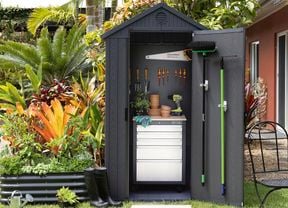 A small dark grey shed with its door open to show a drawer cabinet, tools on a pegboard and a rake and broom hanging on the door