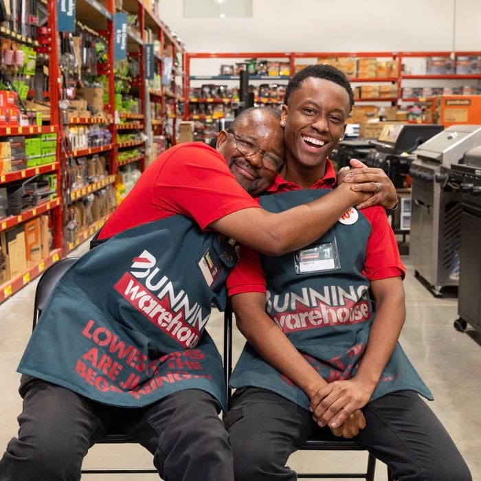 wo Bunnings team members seated in a store aisle, wearing red shirts and green Bunnings aprons, with one team member embracing the other. Shelves of tools and barbecue displays are visible in the background.