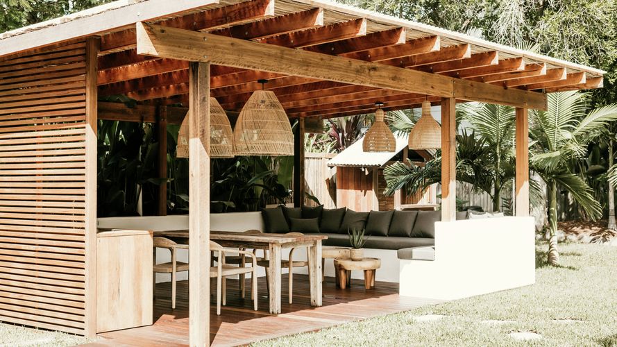 Timber-covered outdoor pavilion with a dining table, built-in lounge seating and woven pendant lights, surrounded by tropical gardens and a lawn.