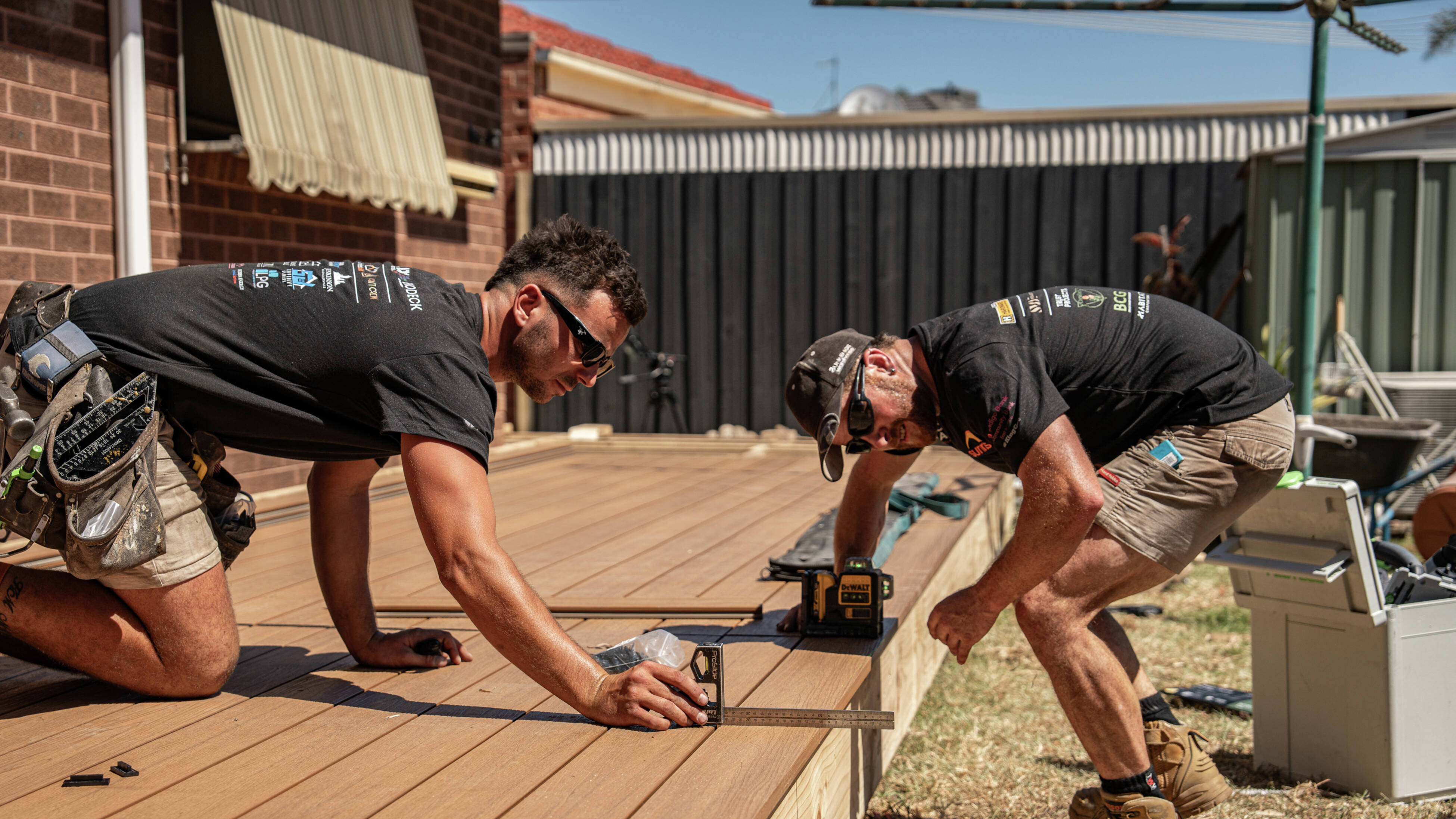 Two tradies wearing sunglasses are crouching over a deck they have built using composite decking materials