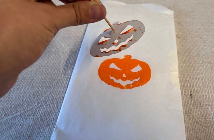 A hand lifts the stamp to reveal a pumpkin in orange paint on a white lolly bag