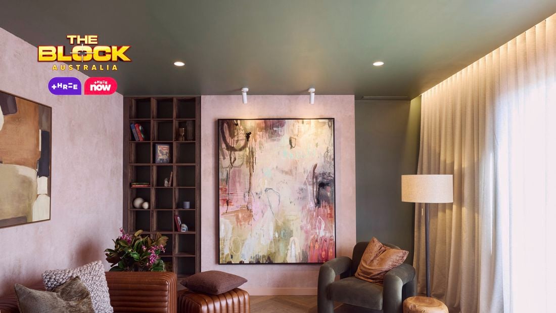 A modern living room featuring a large abstract painting on the wall, a brown leather sofa with cushions, an armchair, a wooden side table, and a floor lamp. A bookshelf with books and decorative items stands nearby. The room has soft lighting and curtains covering the windows. 