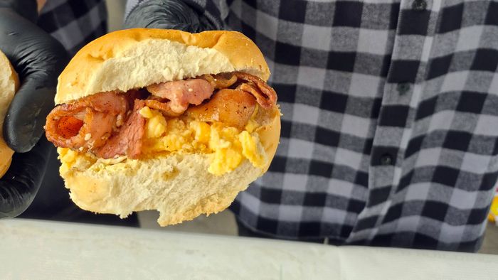 Gloved hands hold a breakfast roll filled with bacon and scrambled egg. The person serving the roll wears a black-and-grey check flannel shirt with a Movember logo on the chest.