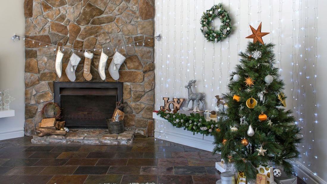 Christmas tree set up in living room with stone fireplace. Fairy lights hang across the wall and various other decorations are placed around the room.