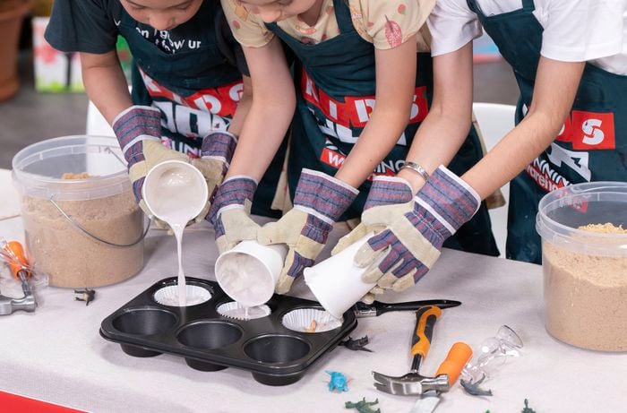 Three kids wearing gloves and aprons are pouring plaster of paris mixture into the moulds to  make the fossil in Bunnings D.I.Y. workshop.