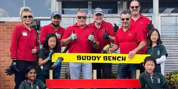 Many Bunnings Team Member's and children in front of a bench with writing on it saying buddy bench.
