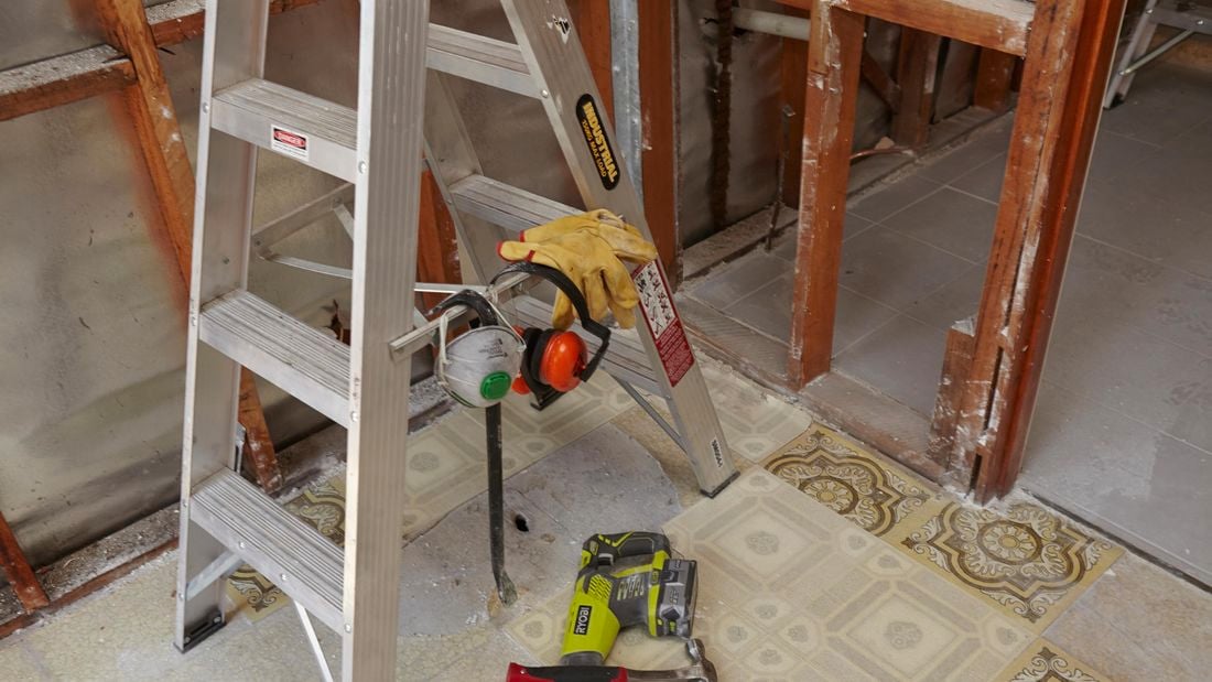 A ladder set up in front of a stud wall, with a pry bar and safety equipment hung from the spreaders and a nearby hammer and nail gun on the floor