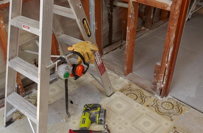 A ladder set up in front of a stud wall, with a pry bar and safety equipment hung from the spreaders and a nearby hammer and nail gun on the floor