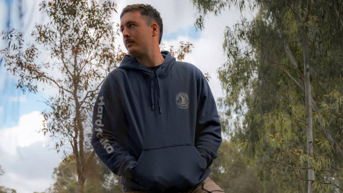 Person standing outdoors in a wooded area with one foot on a tree stump, wearing a hoodie and work pants, with a chainsaw on the ground nearby.