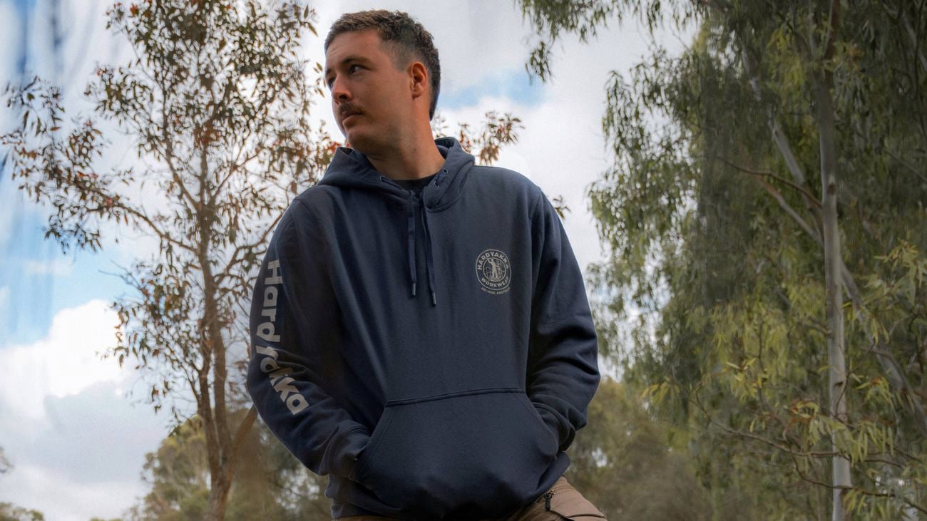 Person standing outdoors in a wooded area with one foot on a tree stump, wearing a hoodie and work pants, with a chainsaw on the ground nearby.